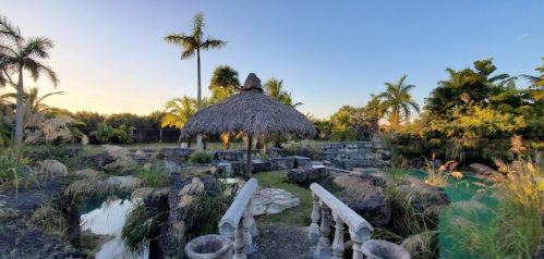 A serene outdoor scene featuring a thatched gazebo surrounded by lush greenery and stone pathways at sunset.