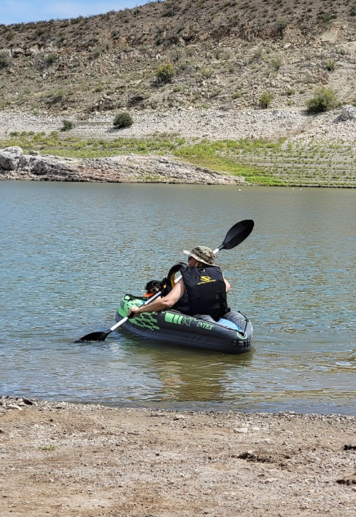 A person in a kayak paddles on a calm lake with a dog sitting beside them, surrounded by rocky terrain.