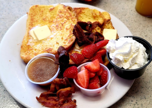 A plate of French toast with butter, bacon, strawberries, whipped cream, and syrup in small cups.