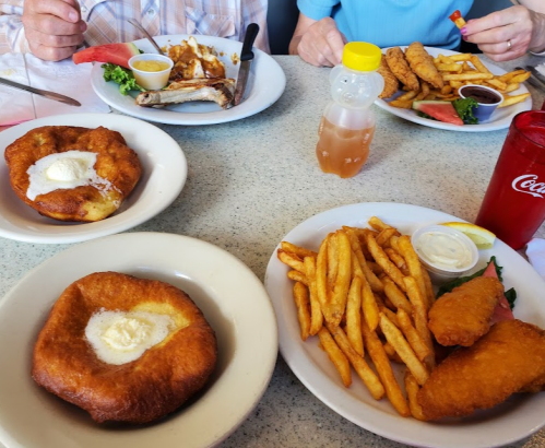 A table with two plates of food: fried fish, fries, and two desserts topped with cream, alongside drinks.