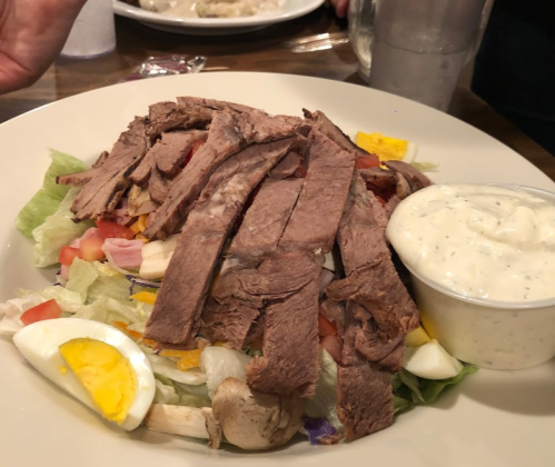 A salad topped with sliced beef, hard-boiled egg, tomatoes, and a side of creamy dressing.