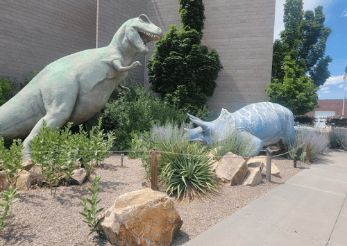Two large dinosaur sculptures, a T-Rex and a triceratops, surrounded by greenery and rocks in a sunny outdoor setting.