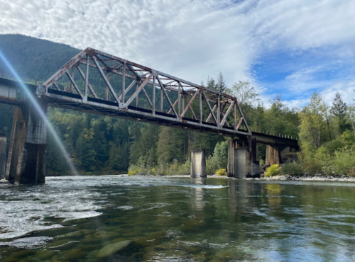 A weathered metal bridge spans a calm river, surrounded by lush green trees and mountains under a partly cloudy sky.