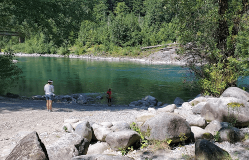 A serene river scene with a person walking on the shore and another person with a dog wading in the water.