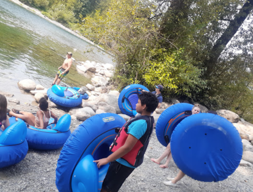 A group of children and adults prepare to go tubing on a river, holding blue inner tubes near rocky shore.