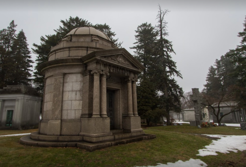 A stone mausoleum with a domed roof, surrounded by trees and patches of snow in a foggy cemetery setting.