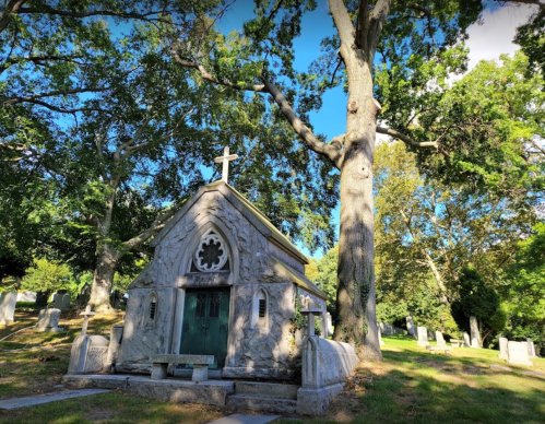 A stone mausoleum with a cross, surrounded by trees in a serene cemetery setting.