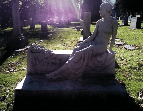 A serene stone statue of a seated woman on a grave, surrounded by a peaceful cemetery landscape.