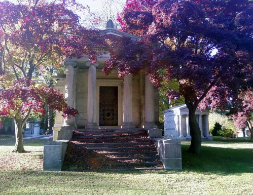 A stone mausoleum with columns surrounded by colorful autumn trees and fallen leaves on the ground.