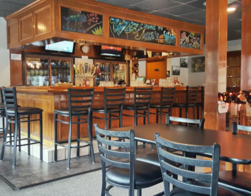 A cozy bar area with wooden accents, high stools, and a chalkboard welcoming guests. Tables are set for dining.