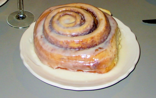 A large, glazed cinnamon roll on a white plate, showcasing its spiral shape and sticky icing.