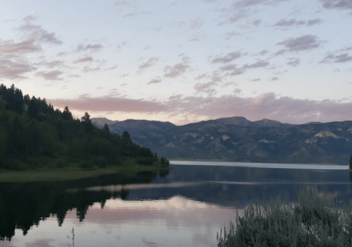A serene lake at sunset, surrounded by mountains and trees, with soft clouds reflecting on the water's surface.