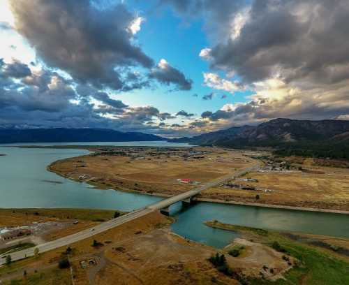 Aerial view of a landscape with a bridge over a river, fields, and mountains under a cloudy sky.