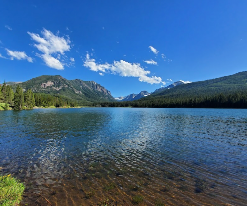 A serene lake surrounded by mountains and lush greenery under a clear blue sky with scattered clouds.