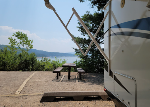 A camper parked by a picnic area with a view of a lake and trees under a clear blue sky.