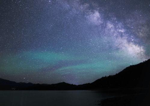 A starry night sky with the Milky Way and green auroras over a calm lake and silhouetted mountains.