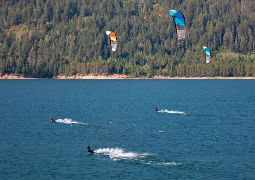 Four kite surfers glide across a blue lake, surrounded by green forested hills under a clear sky.