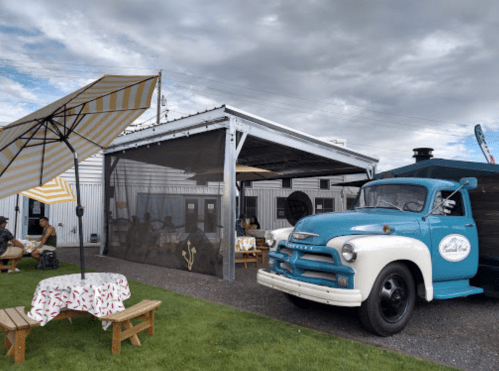 A vintage blue truck parked near a shaded outdoor seating area with picnic tables and umbrellas.