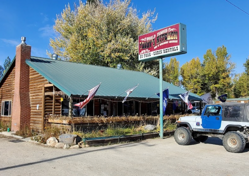 A rustic log cabin building with a green roof, flags, and a sign for rentals, surrounded by trees and gravel.