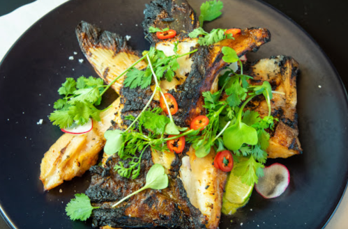 Grilled fish fillets garnished with fresh herbs, chili slices, and lime on a black plate.