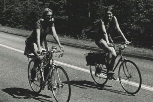 Two women riding bicycles on a quiet road, surrounded by trees, in a black and white photograph.