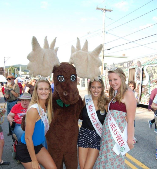 Three young women in sashes pose with a large moose mascot at a festive outdoor event.