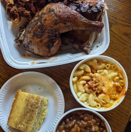 A takeout meal featuring grilled chicken, macaroni and cheese, baked beans, cornbread, and dried flowers on a wooden table.