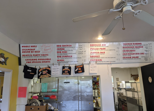 Menu board displaying various BBQ dishes and prices in a casual dining setting. Kitchen visible in the background.