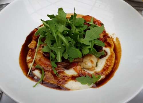 A plate of creamy polenta topped with shrimp, vegetables, and fresh arugula, drizzled with a dark sauce.