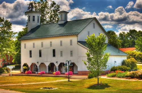 A white barn with a green roof, surrounded by trees and colorful flowers under a partly cloudy sky.