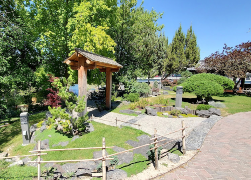 A serene garden featuring a wooden gazebo, lush greenery, rocks, and a winding path under a clear blue sky.