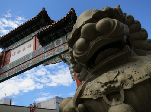A stone lion statue in front of a colorful archway with Chinese characters against a blue sky.