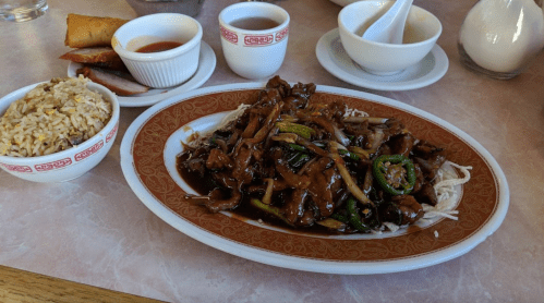 A plate of beef stir-fry with vegetables, served with rice, spring rolls, and dipping sauces on a table.