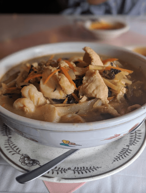 A bowl of chicken soup with vegetables, served on a decorative plate with a spoon beside it.