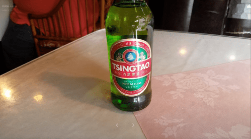 A green bottle of Tsingtao beer sits on a table with a floral tablecloth.