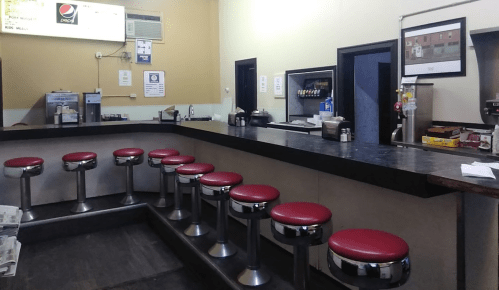 A retro diner interior featuring a long counter with red stools and a menu board on the wall.