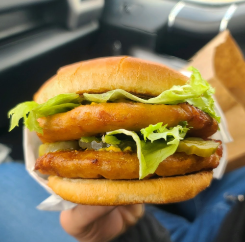 A hand holding a sandwich with two crispy patties, lettuce, and pickles on a soft bun.