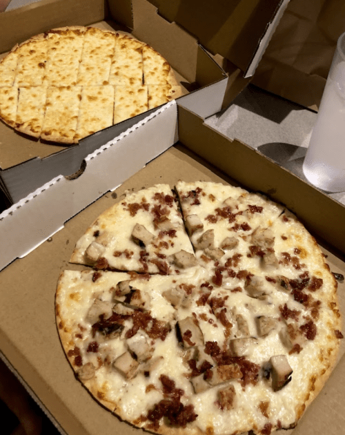 A pizza box containing a large pizza with chicken and bacon, alongside a smaller cheese pizza.