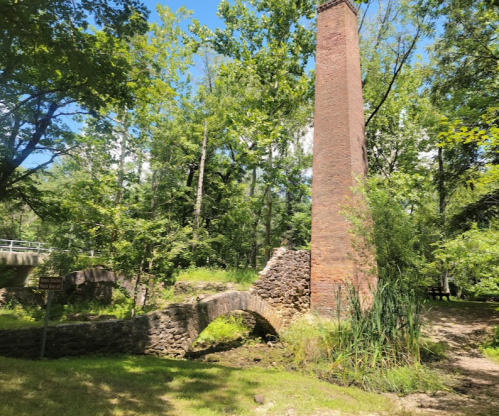 A tall brick chimney stands among lush green trees, next to a stone arch bridge over a small stream.