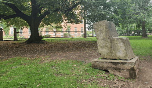 A weathered stone sculpture sits on a grassy area under a large tree, with a building visible in the background.