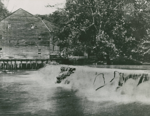 Black and white photo of a wooden mill beside a river, with a waterfall and trees in the background.