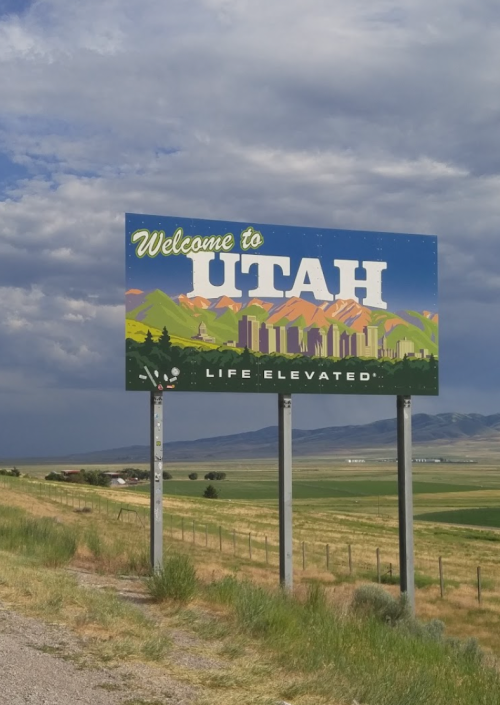 A colorful "Welcome to Utah" sign with mountains and fields under a cloudy sky. Text reads "Life Elevated."