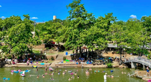 A sunny river scene with people swimming and relaxing on the shore, surrounded by trees and a bridge in the background.