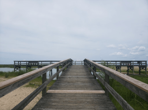 A wooden walkway leads to a calm waterway, surrounded by grass and under a cloudy sky.