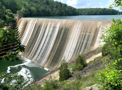 A large dam with cascading water, surrounded by lush green trees and a calm lake under a blue sky.