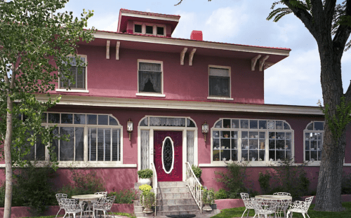 A charming pink house with large windows, a red door, and outdoor seating surrounded by greenery.
