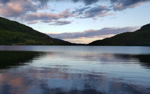 A serene lake surrounded by green hills under a cloudy sky, reflecting the landscape in calm waters.
