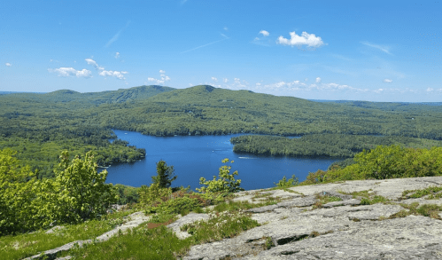 A scenic view of a lake surrounded by lush green hills under a clear blue sky with scattered clouds.
