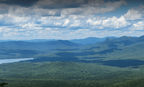 A panoramic view of lush green mountains and a lake under a partly cloudy sky.