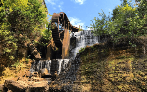 A waterwheel beside a cascading waterfall, surrounded by lush greenery and a blue sky.